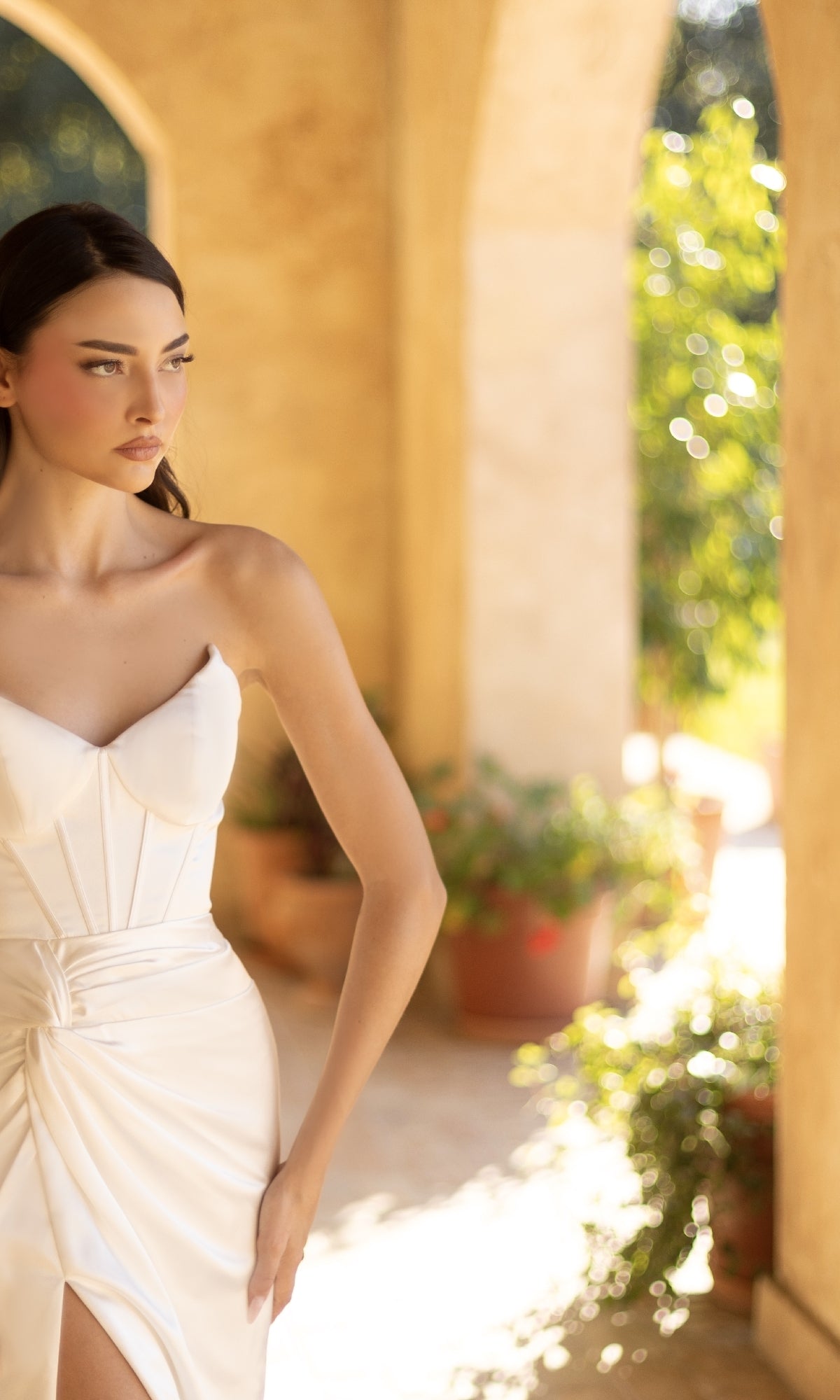 A woman in an Off-White Strapless Prom Gown (Tina Holly TT003W) stands in a sunlit corridor with arched openings and potted plants in the background.